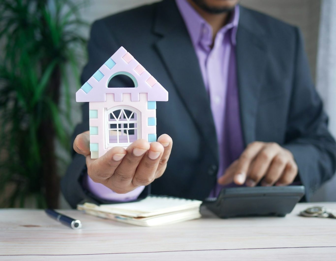 Hombre en traje sosteniendo una casa de juguete y usando una calculadora.