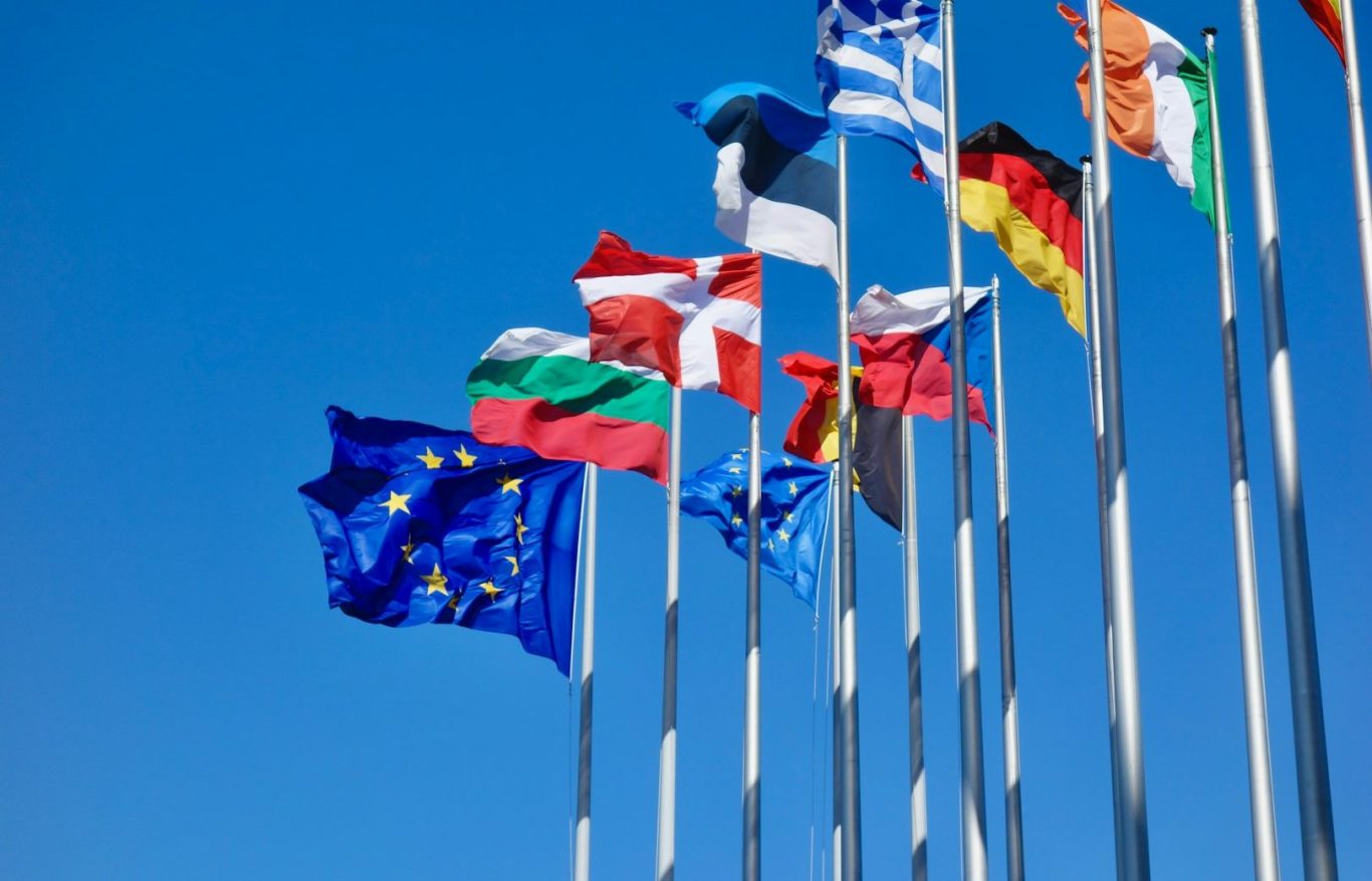 Varios banderas ondeando contra un cielo azul claro, incluyendo la de la Unión Europea.