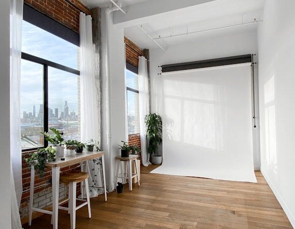 Espacio luminoso con pared blanca, plantas y mesa, vista urbana al fondo.