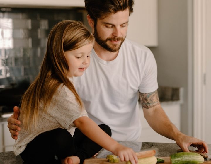 Padre e hija preparando alimentos juntos en la cocina. La niña observa atentamente.
